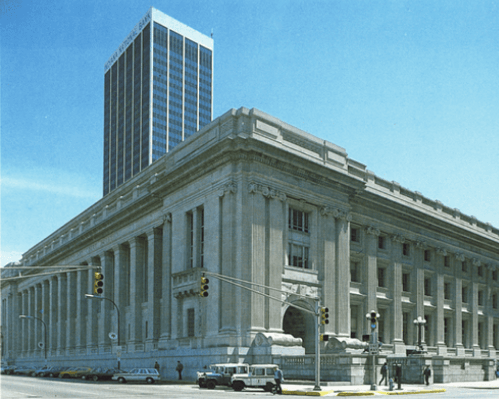Untitled-design-2 A historic, large concrete building with large columns stands on a street corner with modern skyscrapers in the background and a few cars and pedestrians at the intersection.